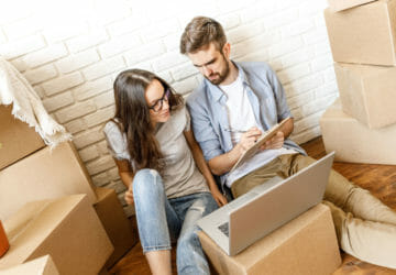 Young casual woman and man sitting on floor with carton boxes and laptop taking notes on clipboard and talking.