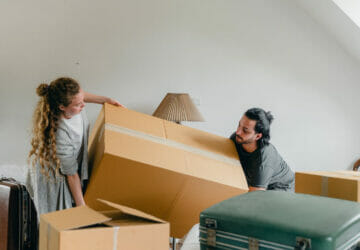 A man and a woman struggle to lift a heavy moving box in their living room