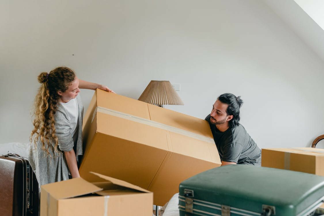 A man and a woman struggle to lift a heavy moving box in their living room