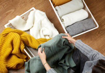 Person folding winter sweaters on a wooden floor beside an organizer bin with neatly folded clothing.