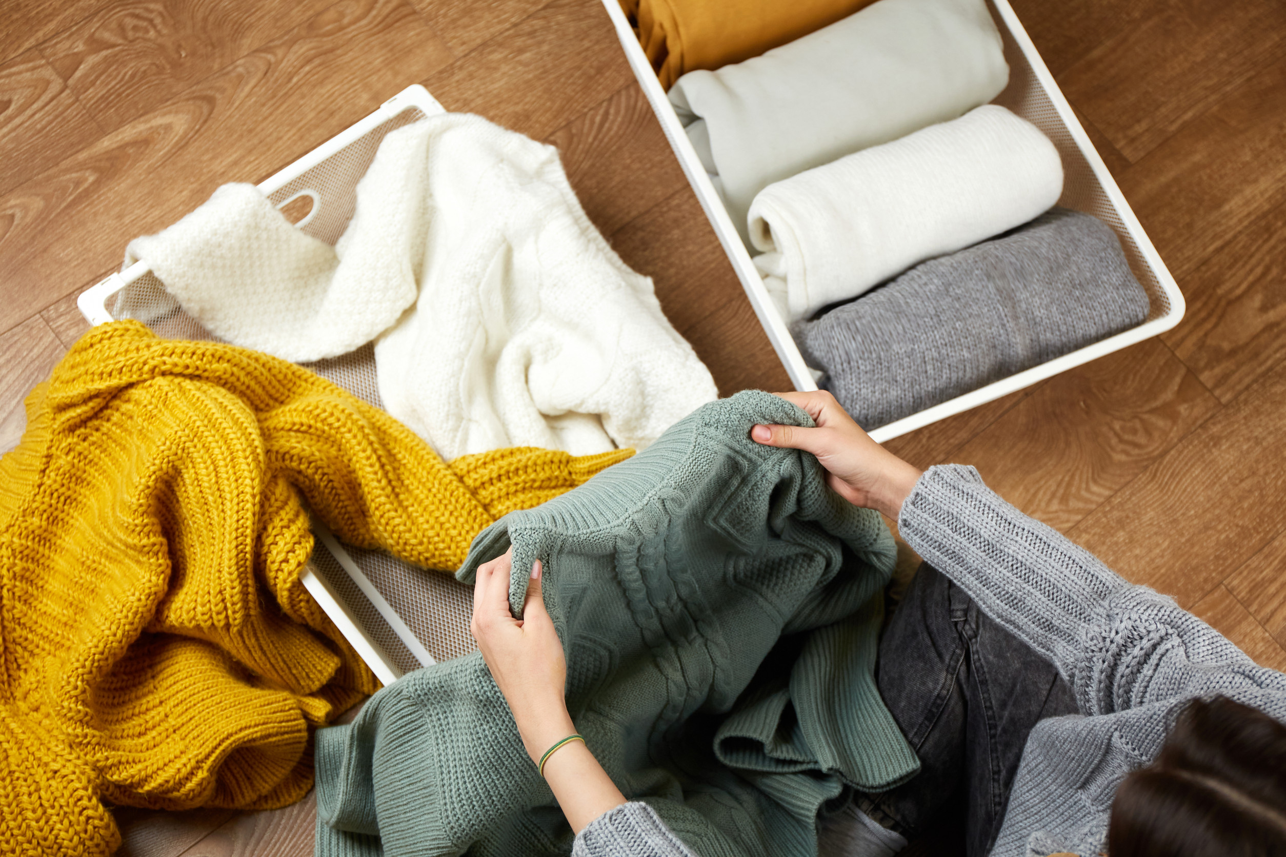 Person folding winter sweaters on a wooden floor beside an organizer bin with neatly folded clothing.
