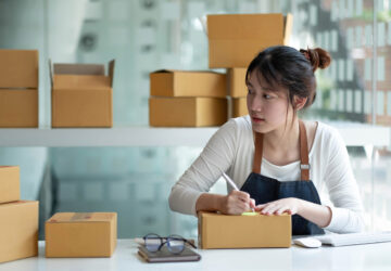 Woman packing and labeling shipping boxes for a small business order at a home workspace