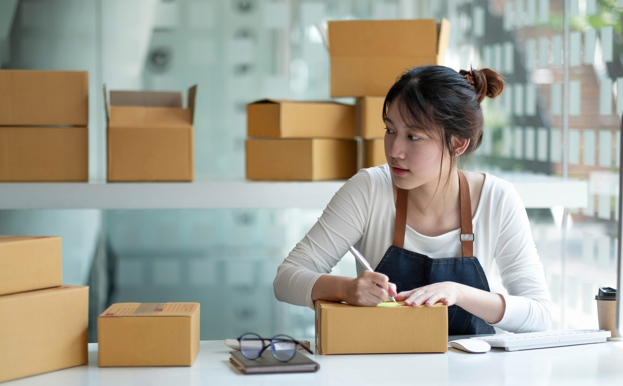 Woman packing and labeling shipping boxes for a small business order at a home workspace