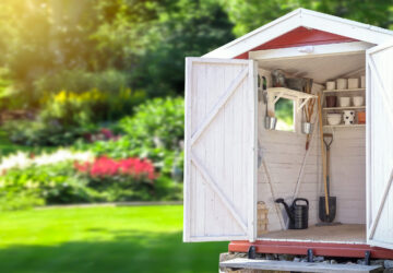 Abri de jardin ouvert avec étagères organisées, outils de jardinage et arrosoir dans une cour arrière paysagée et ensoleillée.