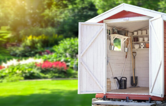 Abri de jardin ouvert avec étagères organisées, outils de jardinage et arrosoir dans une cour arrière paysagée et ensoleillée.