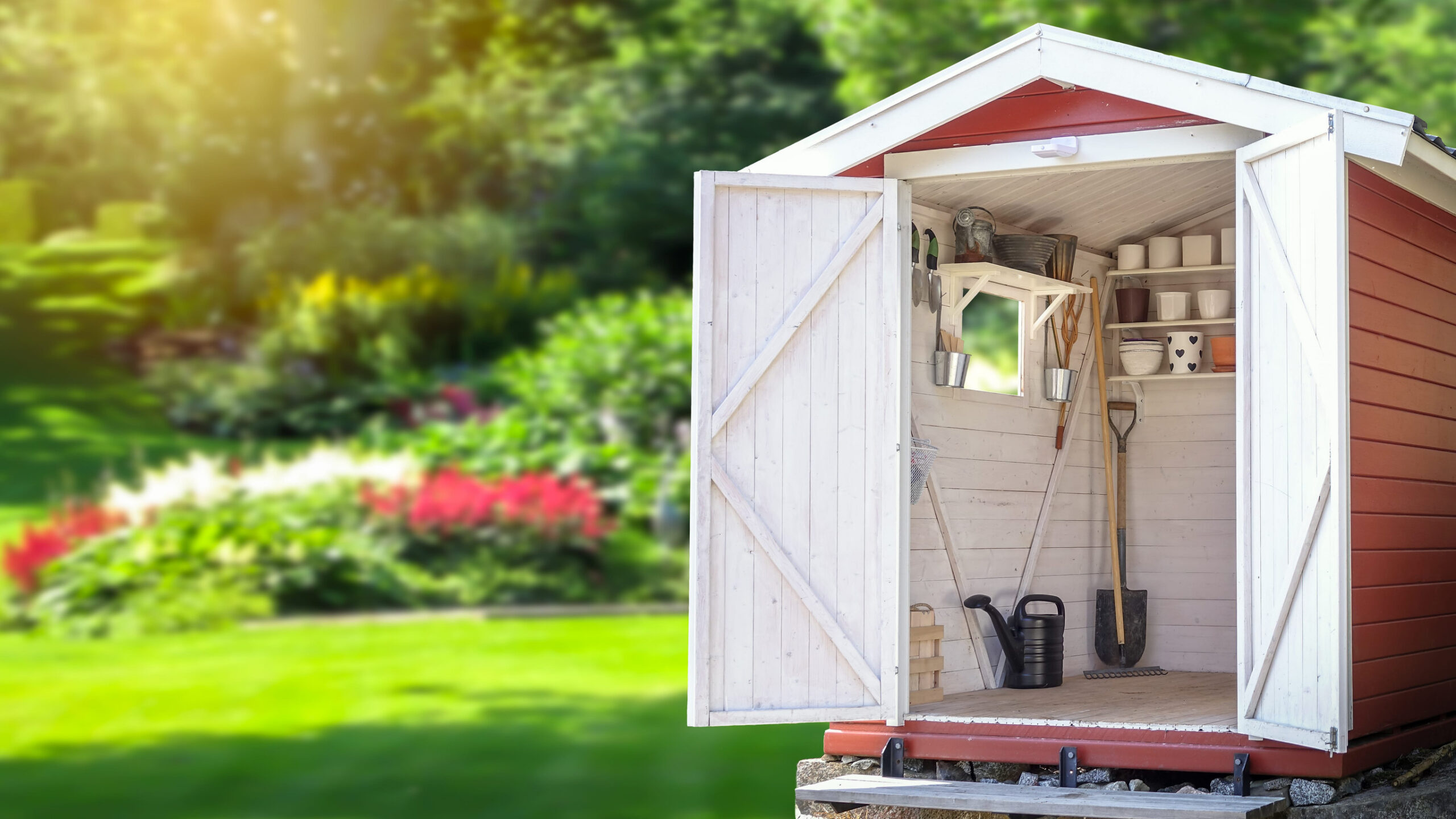 Open backyard garden shed with organized shelves, gardening tools, and watering can in a sunny landscaped garden.