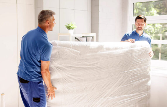 Two movers carrying a mattress wrapped in plastic for storage