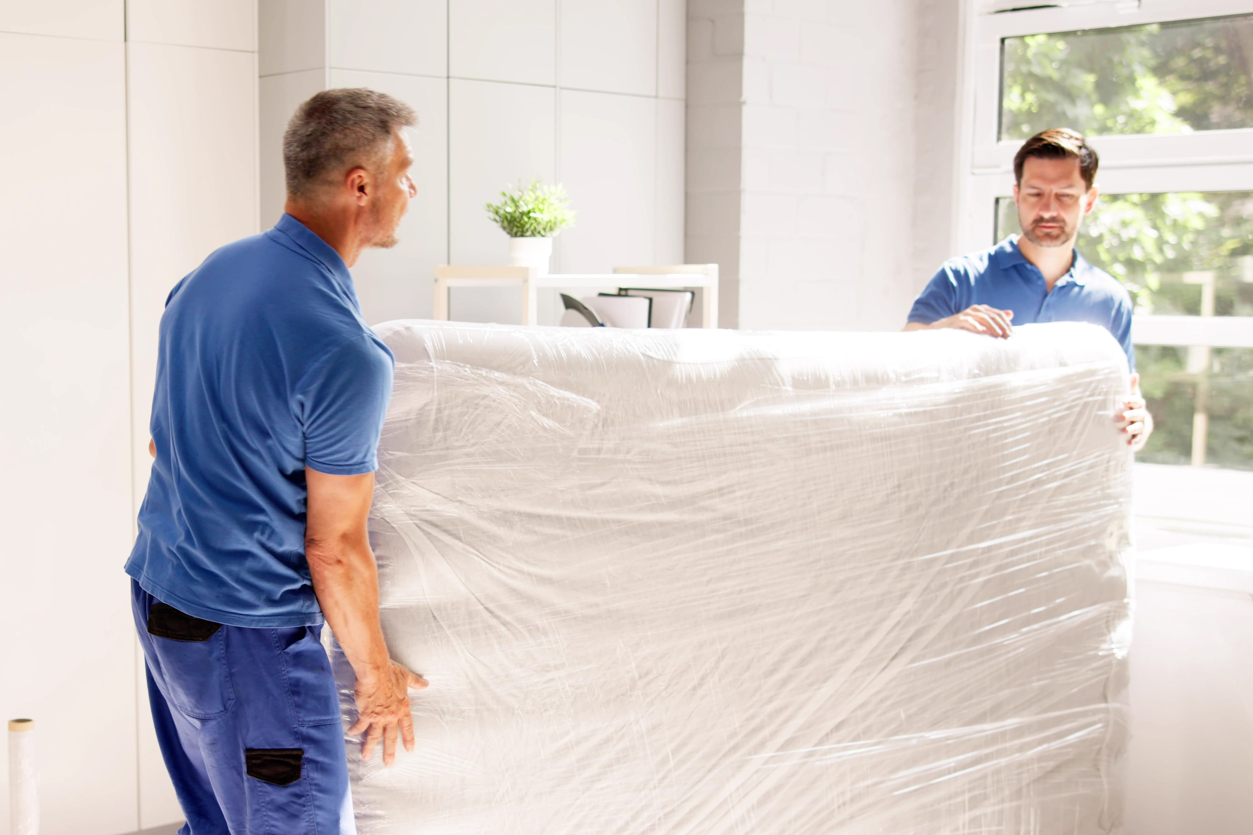 Two movers carrying a mattress wrapped in plastic for storage